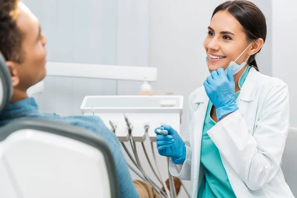 Alegre dentista femenina sosteniendo máscara y taladro y sonriendo cerca del paciente - foto de stock