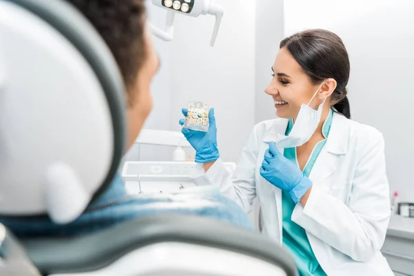 Estomatóloga sonriente mirando el modelo de mandíbula dental con frenos cerca del paciente - foto de stock