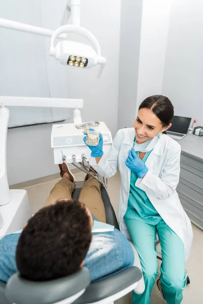 Улыбающаяся женщина-стоматолог показывает модель челюсти с брекетами к пациенту в кресле — стоковое фото
