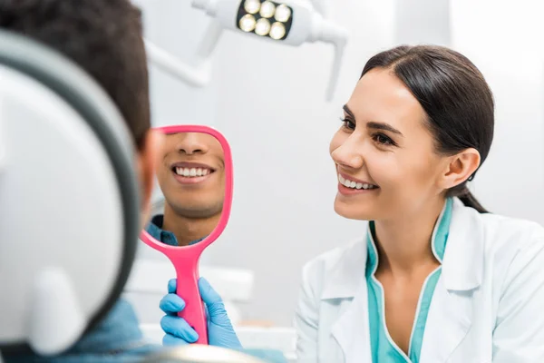 Bonito afro-americano homem sorrindo enquanto atraente dentista segurando espelho — Fotografia de Stock