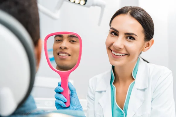 Guapo afroamericano hombre sonriendo mientras mujer dentista sosteniendo espejo - foto de stock