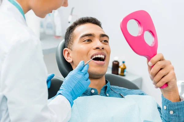 Dentista mujer sosteniendo instrumento médico cerca de paciente afroamericano mirando el espejo - foto de stock