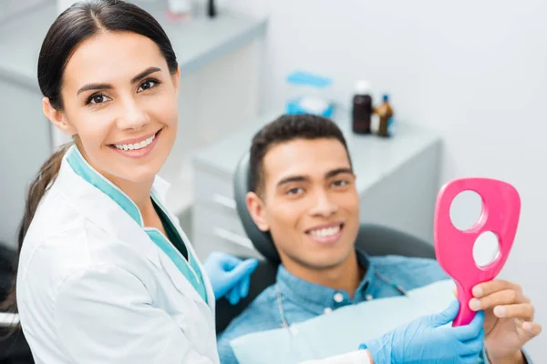 Enfoque selectivo de la atractiva dentista femenina de pie cerca de un paciente afroamericano - foto de stock