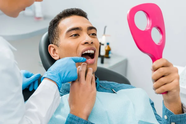 Dentiste femelle touchant dent de bel homme afro-américain regardant miroir — Photo de stock