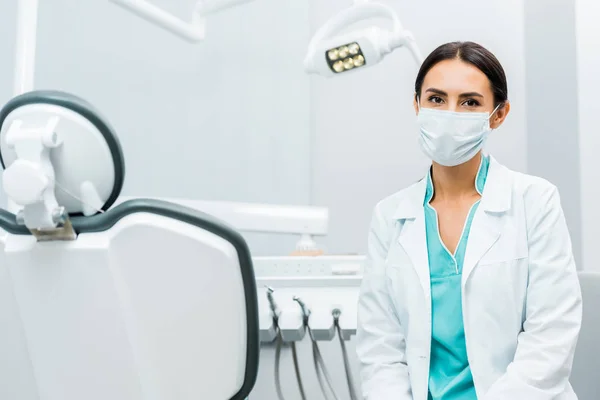 Female dentist in white coat and mask near dental chair — Stock Photo