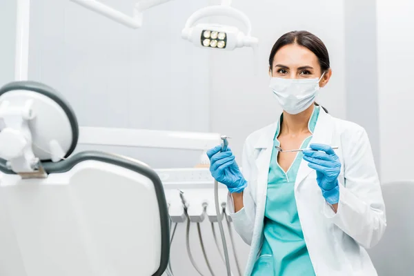Female dentist in white coat and mask holding dental instruments — Stock Photo