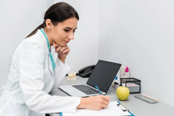 Médico femenino reflexivo haciendo notas cerca de la computadora portátil - foto de stock