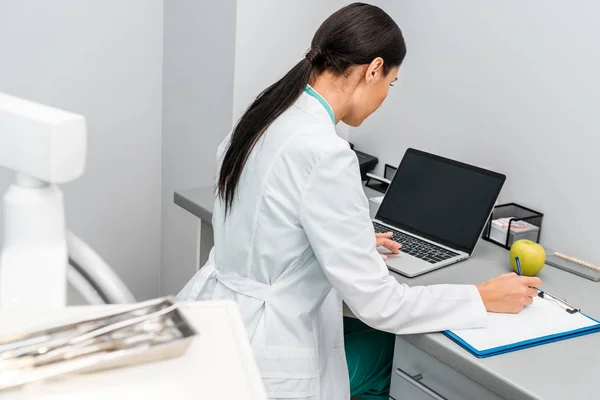 Ärztin benutzt Laptop mit leerem Bildschirm und macht sich Notizen — Stockfoto