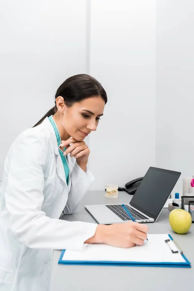 Nachdenkliche Ärztin macht sich Notizen in Laptopnähe — Stockfoto