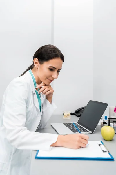 Pensive female doctor making notes near laptop — Stock Photo