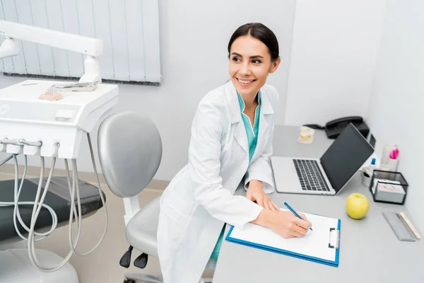 Attractive female doctor making notes near laptop — Stock Photo