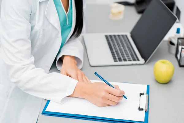 Vista recortada de la doctora haciendo notas cerca de la computadora portátil - foto de stock