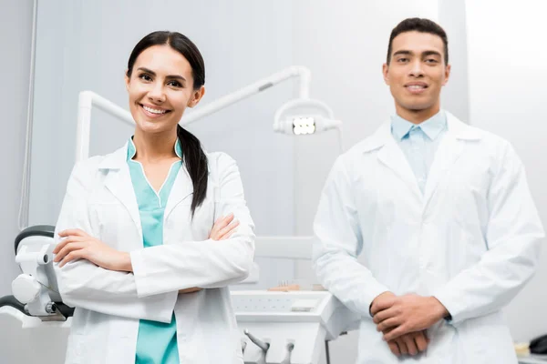 Alegre médico feminino em pé perto de colega de trabalho afro-americano em casaco branco com braços cruzados — Fotografia de Stock