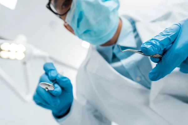 Селективная направленность стоматологических инструментов в руках мужчины-стоматолога в очках и маске — стоковое фото