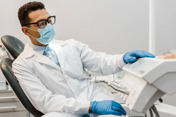Dentista afroamericano sentado en gafas, máscara y abrigo blanco - foto de stock