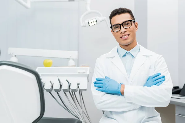 Guapo afroamericano dentista en gafas de pie con los brazos cruzados - foto de stock