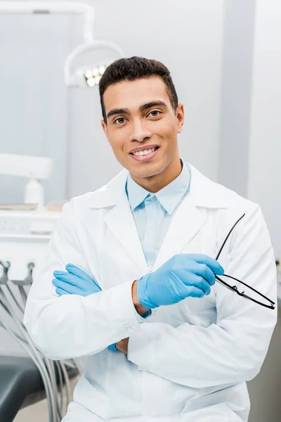 Apuesto dentista afroamericano en gafas de pie con los brazos cruzados y sosteniendo gafas - foto de stock