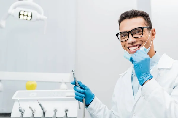 Sonriente dentista afroamericano en gafas sosteniendo taladro - foto de stock