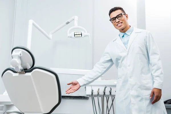 Sonriente afroamericano médico mostrando silla en clínica dental - foto de stock