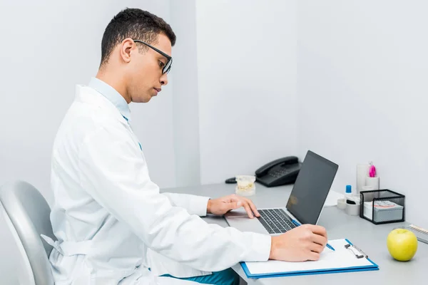 Handsome african american doctor making notes and using laptop — Stock Photo