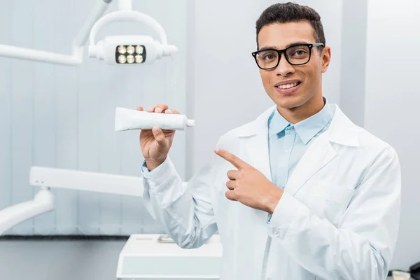 Handsome african american doctor in glasses pointing at toothpaste in dental clinic — Stock Photo