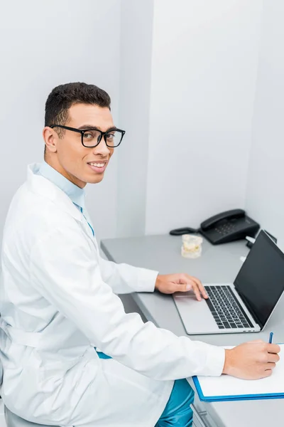 Alegre afroamericano médico toma notas y el uso de ordenador portátil - foto de stock