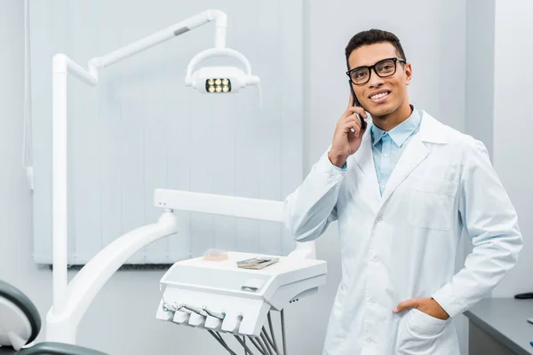 Médecin afro-américain souriant en lunettes et manteau blanc parlant sur smartphone avec la main dans la poche — Photo de stock