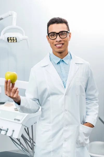 Handsome african american dentist with hand in pocket holding apple — Stock Photo