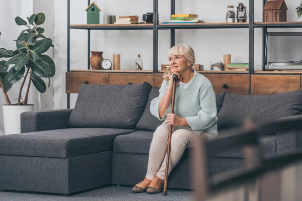 Dreamy senior woman sitting on sofa with walking cane in living room — Stock Photo