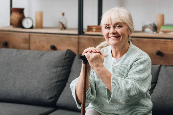 Cheerful senior woman sitting on sofa and holding walking stick — Stock Photo