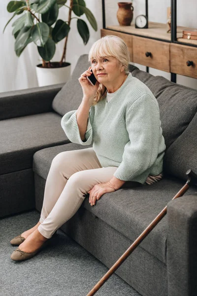 Sad senior woman talking on smartphone while sitting on sofa — Stock Photo