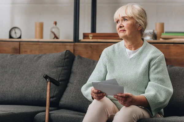 Verärgerte Rentnerin mit blonden Haaren hält alte Fotos hoch, während sie auf Sofa sitzt — Stockfoto