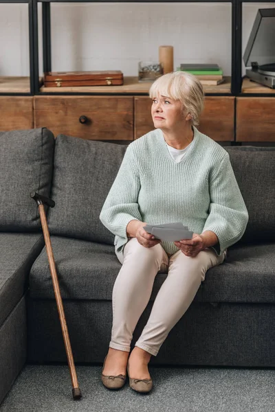 Verärgerte Rentnerin mit blonden Haaren hält Fotos hoch, während sie im Wohnzimmer auf dem Sofa sitzt — Stockfoto