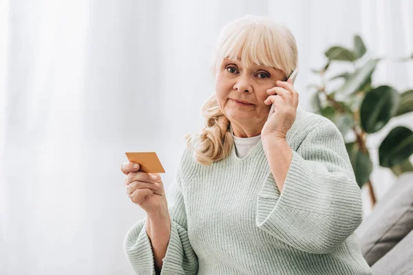 Mulher aposentada confuso segurando cartão de crédito ao falar no smartphone — Fotografia de Stock
