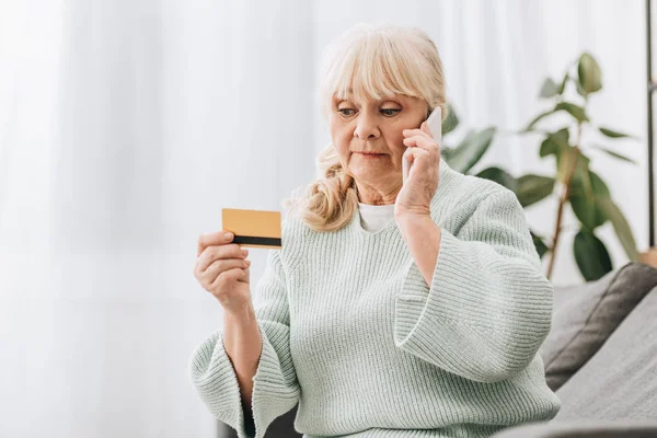 Mujer mayor confundida mirando la tarjeta de crédito mientras habla en el teléfono inteligente - foto de stock