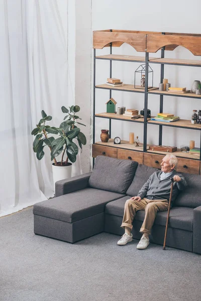 Retired man with grey hair holding walking cane while sitting on sofa — Stock Photo