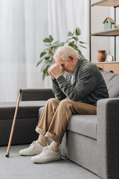 Sconvolto pensionato maschio seduto sul divano vicino a bastone da passeggio — Foto stock