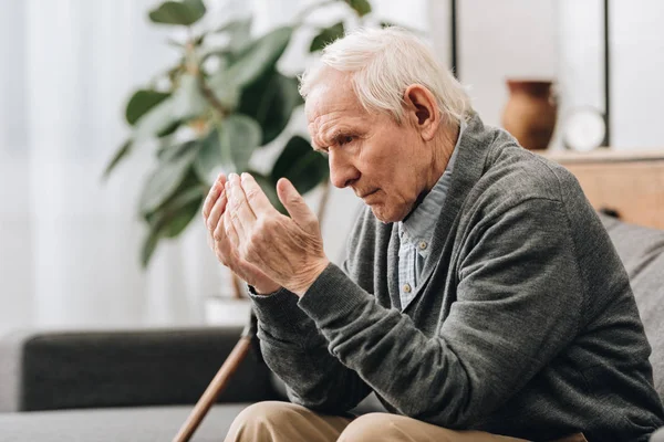 Пенсионер с седыми волосами смотрит на руки, сидя на диване — стоковое фото