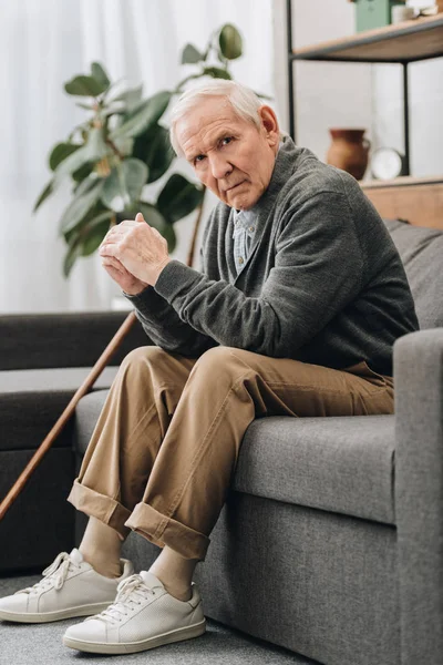 Upset retired man sitting on sofa near walking stick in living room — Stock Photo