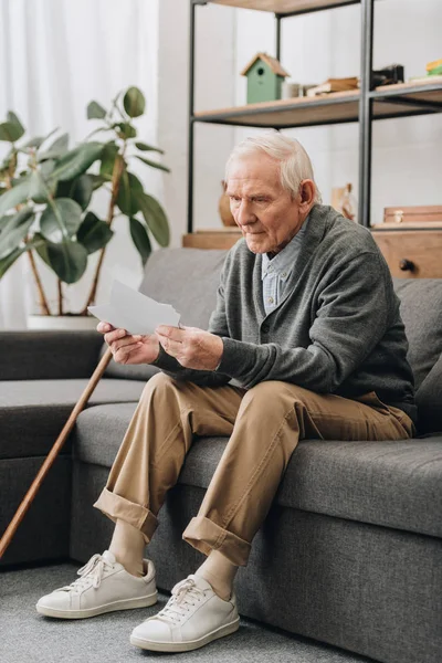 Pensionista triste con el pelo gris mirando fotos mientras está sentado en el sofá - foto de stock