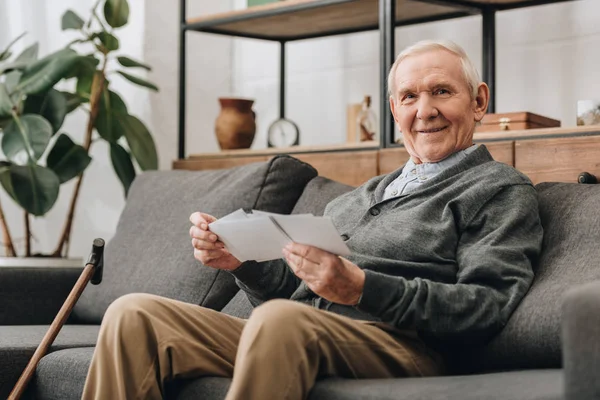 Alegre hombre mayor con pelo gris sosteniendo fotos y sentado en el sofá - foto de stock