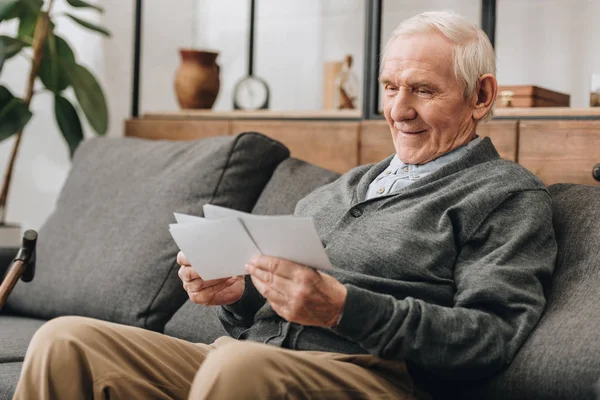 Glücklicher älterer Mann mit grauen Haaren schaut sich Fotos an und sitzt auf dem Sofa — Stockfoto