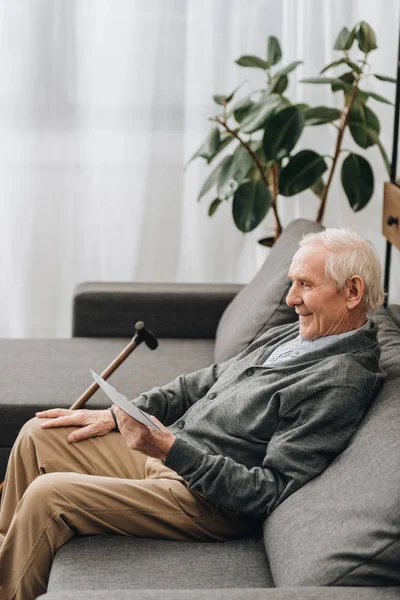 Улыбающийся пенсионер с седыми волосами, смотрящий на фотографии и сидящий на диване — стоковое фото