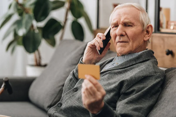 Hombre mayor mirando la tarjeta de crédito mientras habla en el teléfono inteligente - foto de stock