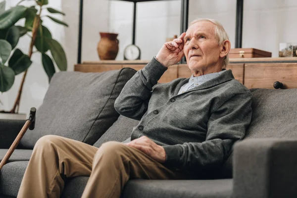 Hombre mayor reflexivo con el pelo gris sentado en el sofá en la sala de estar - foto de stock