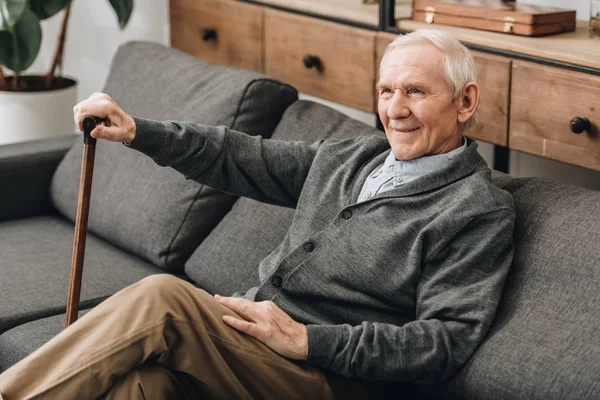 Улыбающийся пенсионер, сидящий на диване с тростью — стоковое фото