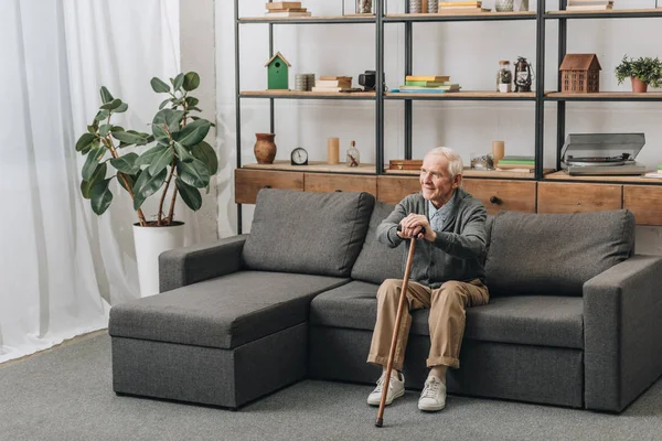Glücklicher älterer Mann lächelt und hält Gehstock, während er auf dem Sofa sitzt — Stockfoto