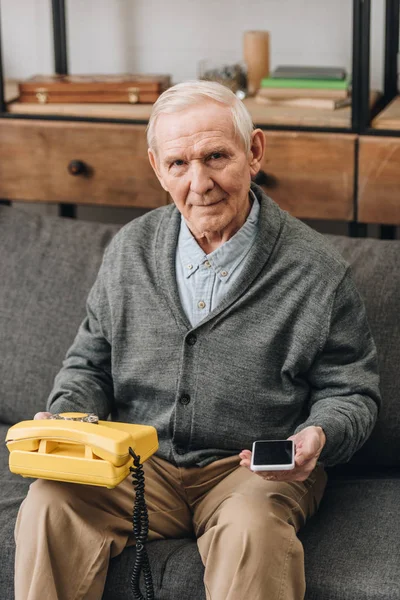 Aposentado homem segurando smartphone e telefone retro enquanto sentado no sofá — Fotografia de Stock