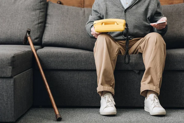 Vista cortada do homem aposentado segurando smartphone e telefone retro enquanto sentado no sofá — Fotografia de Stock