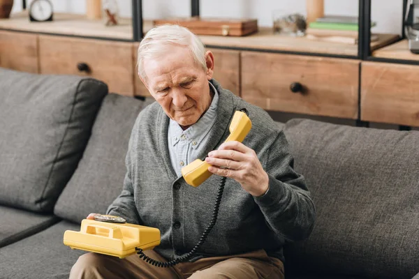 Rentner schaut auf altes Handy, während er auf Sofa sitzt — Stockfoto